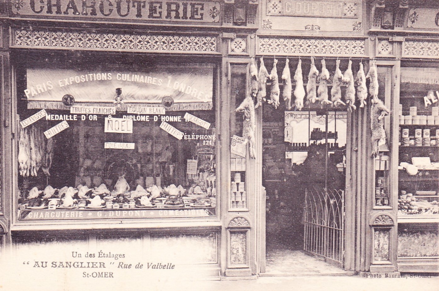 Ce commerce de charcuterie, chez M. G Dupont est bien achalandée. On peut y voir les poulets dans la vitrine et les lapins qui pendent au dessus de la porte. 